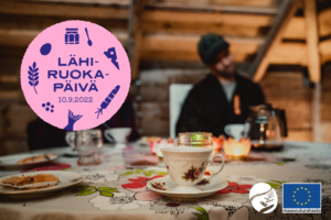 Lähiruokapäivä mainoskuva, jossa ruokapöytä ja kahvikuppi. Taustalla ihmisiä.